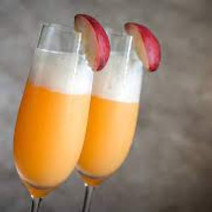 May include: Two champagne flutes filled with an orange-coloured beverage, topped with white foam and garnished with a red apple slice. The glasses are angled, creating a visually appealing composition. The background is a neutral grey.