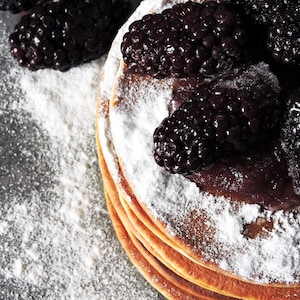 May include: A stack of pancakes topped with blackberries and powdered sugar. The pancakes are golden brown and the blackberries are juicy and plump.