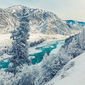 May include: A snowy mountain landscape with a winding turquoise river flowing through it. The river is partially frozen, with ice forming along the edges. The mountains are covered in snow, and the trees are bare and covered in frost.