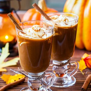 May include: Two glasses of pumpkin spice latte topped with whipped cream and cinnamon sticks. The glasses are on a wooden surface with autumnal decorations, including pumpkins and leaves.