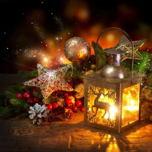 May include: A gold lantern with a deer design and a lit candle inside. The lantern is surrounded by a Christmas tree branch with red berries, a gold star, and a gold bauble ornament. The background is a blurry image of twinkling lights.