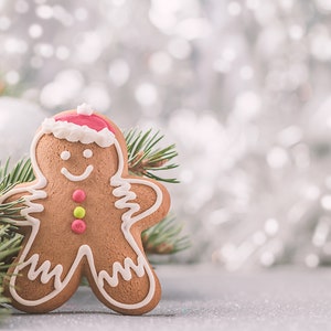 Peut inclure: Un biscuit en pain d'épices en forme d'homme, décoré de glaçage blanc et d'un chapeau de Père Noël rouge et blanc. Le biscuit est posé sur une surface blanche avec une branche de pin verte au premier plan.