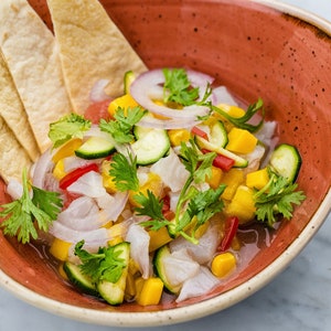 May include: A red bowl filled with a ceviche dish. The ceviche is made with white fish, mango, red onion, coriander, and courgette.  Tortilla chips are on the side.