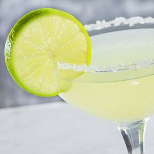 May include: A clear glass margarita glass with a salted rim and a lime wedge garnish. The drink is a pale yellow colour.