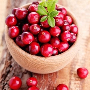 May include: A wooden bowl filled with fresh, red cranberries. The cranberries are arranged in a cluster, with a few green leaves visible on top.