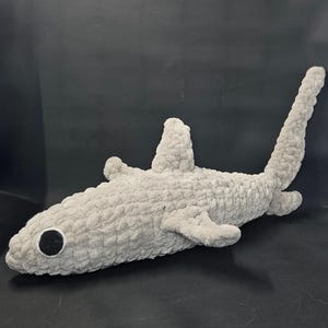 May include: A plush, gray shark toy with a textured, fuzzy surface. The shark has a black eye, fins, and a tail. The toy is designed to resemble a shark.