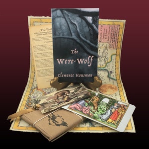 May include: A vintage-style book titled "The Were-Wolf" by Clemence Housman, with a dark forest scene on the cover. The book is displayed on a wooden stand, surrounded by a map, a tarot card, and a small decorative box.