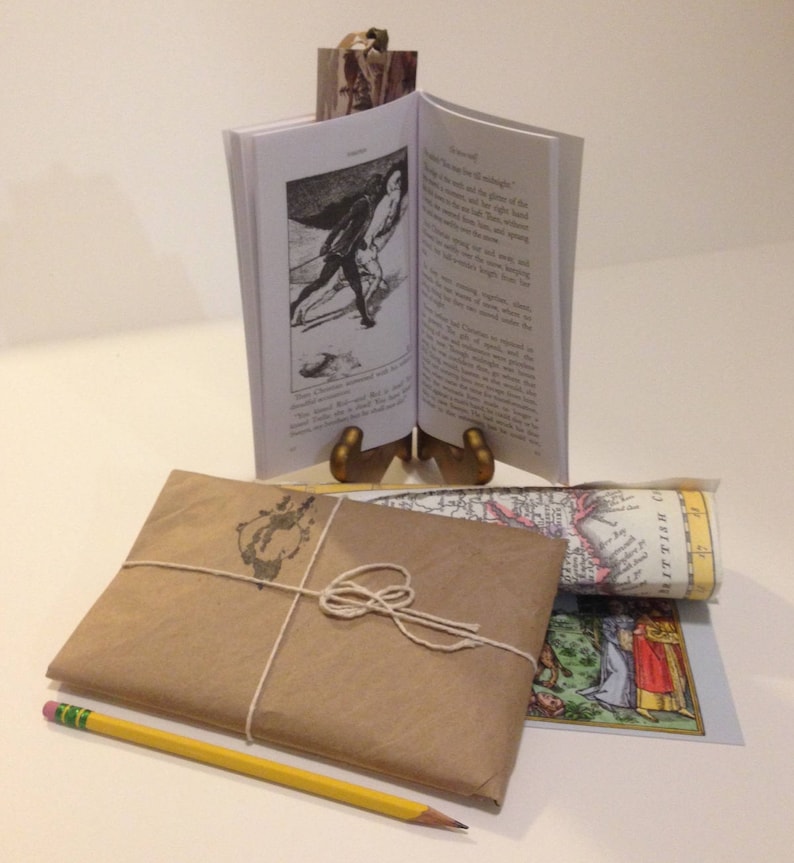 May include: A brown paper package tied with twine, a yellow pencil, and a rolled up map with the text "WESTPHALIA" visible. A small book with a black and white illustration of a person in a historical costume is on a gold metal stand.