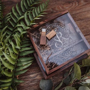May include: A square dark wood box with a clear lid featuring the white engraved text Sophie Rensha Photography and a monogram logo. Inside the box, there is a wooden USB flash drive and its cap resting on a bed of shredded brown paper filler.