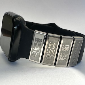 Puede incluir: Apple Watch negro con una banda negra y cuatro dijes de metal plateado. Los dijes muestran el texto "Paris 2025 42KM", "LON 2025 42.2KM", "ATH 2025" y "run junkies".