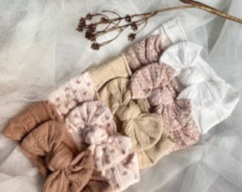 May include: A collection of baby headbands in various shades of beige, pink, and white. The headbands feature different textures and patterns, including knitted and dotted designs. A dried floral stem rests above the headbands on a white, textured surface.