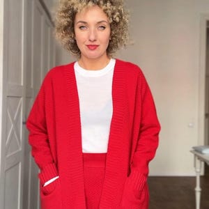 Può includere: Una donna indossa un cardigan rosso acceso con ampie tasche, una gonna abbinata e una maglietta bianca. L'outfit è realizzato in maglia. La donna ha i capelli biondi ricci e sorride.