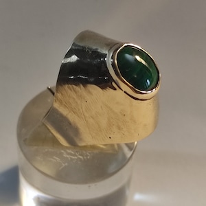 Puede incluir: Un anillo de plata con un engaste de oro que sostiene una gran piedra preciosa ovalada de color verde oscuro. El anillo tiene una banda ancha con una textura martillada. La piedra preciosa es el punto focal, engastada en un bisel de oro.