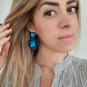 Pode incluir: Um par de brincos azuis e turquesa com uma pedra turquesa grande e facetada e pedras azuis menores. Os brincos são feitos com uma técnica de fio tecido.