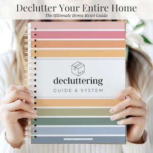 May include: A spiral-bound planner with the text "Declutter Your Entire Home" and "The Ultimate Home Reset Guide." The cover features horizontal stripes in shades of pink, orange, green, and blue. The word "decluttering" is prominently displayed.