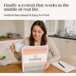Mom sitting at kitchen table with coffee, reading printable decluttering guide and system binder in a real-life home setting with background clutter and notebook beside her