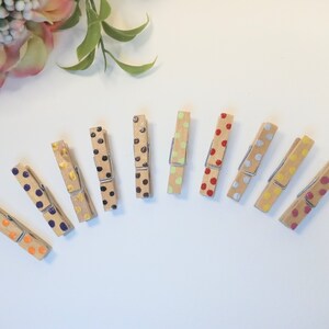 May include: Ten wooden clothespins with colorful polka dots. The clothespins are painted in various colors, including orange, purple, black, gold, brown, green, red, silver, yellow, and pink.