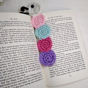 May include: A colorful crocheted bookmark shaped like a caterpillar. The bookmark is made of pink, blue, purple, and white yarn. The bookmark is on top of an open book.