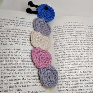 May include: A crocheted bookmark in the shape of a caterpillar with blue, gray, white, and pink crocheted circles. The bookmark is on top of an open book with the text "The Trial in the Circle" visible.