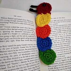 May include: A colorful crocheted caterpillar bookmark with a red head, yellow, orange, blue, and green segments. The bookmark is placed on an open book.