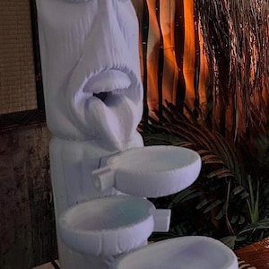 May include: A white, carved tiki statue with an open mouth and tongue, featuring three tiered bowls. The statue is on a patterned surface, with a backdrop of wooden panels and tropical foliage. The overall theme is tropical decor.