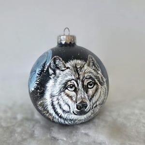 May include: A hand-painted glass Christmas ornament featuring a wolf's face against a black background with a blue moon and white stars.