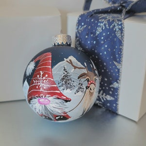 May include: A blue glass Christmas ornament with a hand-painted scene of a gnome and a reindeer in a snowy winter landscape. The gnome is wearing a red hat with white trim and a white beard. The reindeer is brown with a red nose and a red ribbon around its neck.