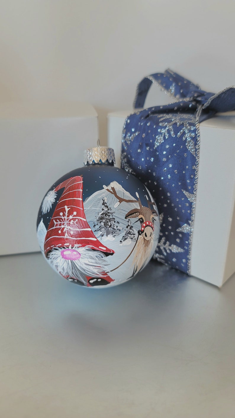 May include: A blue glass Christmas ornament with a hand-painted scene of a gnome and a reindeer in a snowy winter landscape. The gnome is wearing a red hat with white trim and a white beard. The reindeer is brown with a red nose and a red ribbon around its neck.