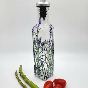 Op de afbeelding: Een heldere glazen fles met een lavendelbloemdessin en een zilveren en zwarte giettuit. De fles staat op een wit oppervlak met twee groene aspergestelen en drie rode tomaten.