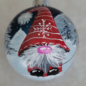 May include: A round Christmas ornament with a painted gnome wearing a red and white hat with a snowflake design. The gnome is standing in a snowy landscape with trees in the background.