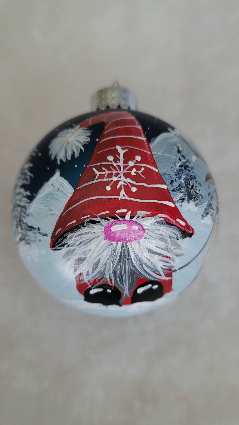 May include: A round Christmas ornament with a painted gnome wearing a red and white hat with a snowflake design. The gnome is standing in a snowy landscape with trees in the background.
