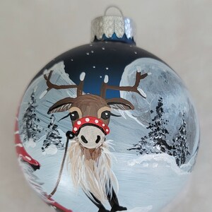 May include: A blue glass Christmas ornament with a hand-painted scene of a reindeer pulling a sleigh through a snowy forest under a full moon.
