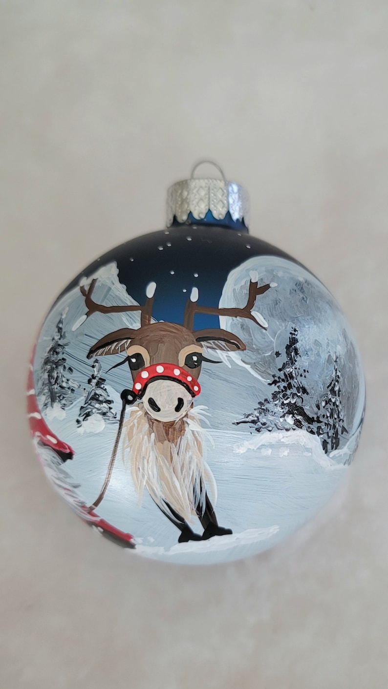 May include: A blue glass Christmas ornament with a hand-painted scene of a reindeer pulling a sleigh through a snowy forest under a full moon.
