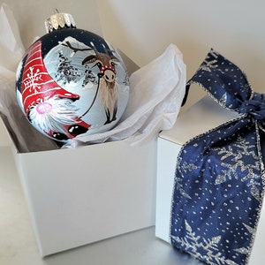 May include: A hand-painted Christmas ornament featuring a reindeer pulling a sleigh with a gnome in a red hat. The ornament is blue with white snow and trees. It is packaged in a white box with a blue ribbon and snowflake design.