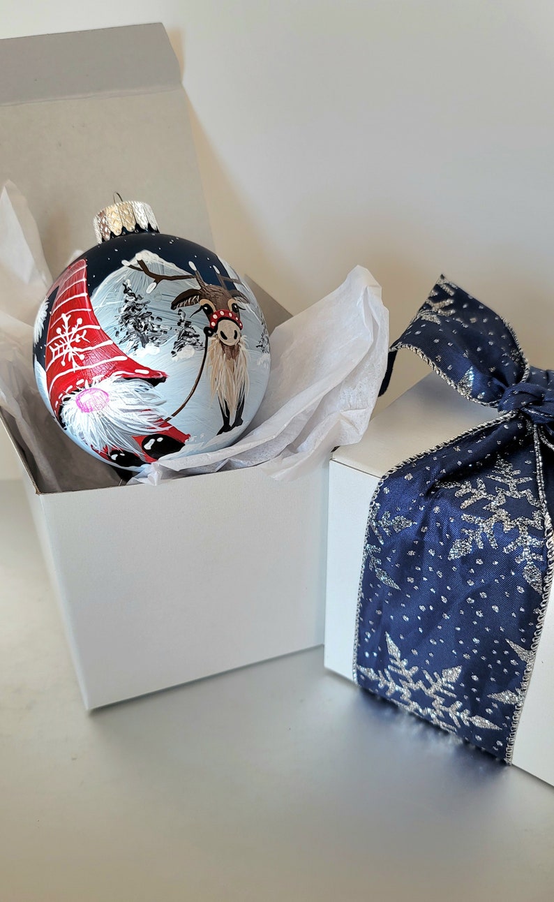 May include: A hand-painted Christmas ornament featuring a reindeer pulling a sleigh with a gnome in a red hat. The ornament is blue with white snow and trees. It is packaged in a white box with a blue ribbon and snowflake design.