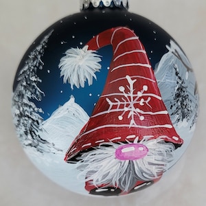 May include: A hand-painted Christmas ornament featuring a gnome with a red and white striped hat, a white beard, and a pink nose. The gnome is set against a blue background with white snow-capped mountains and white trees.
