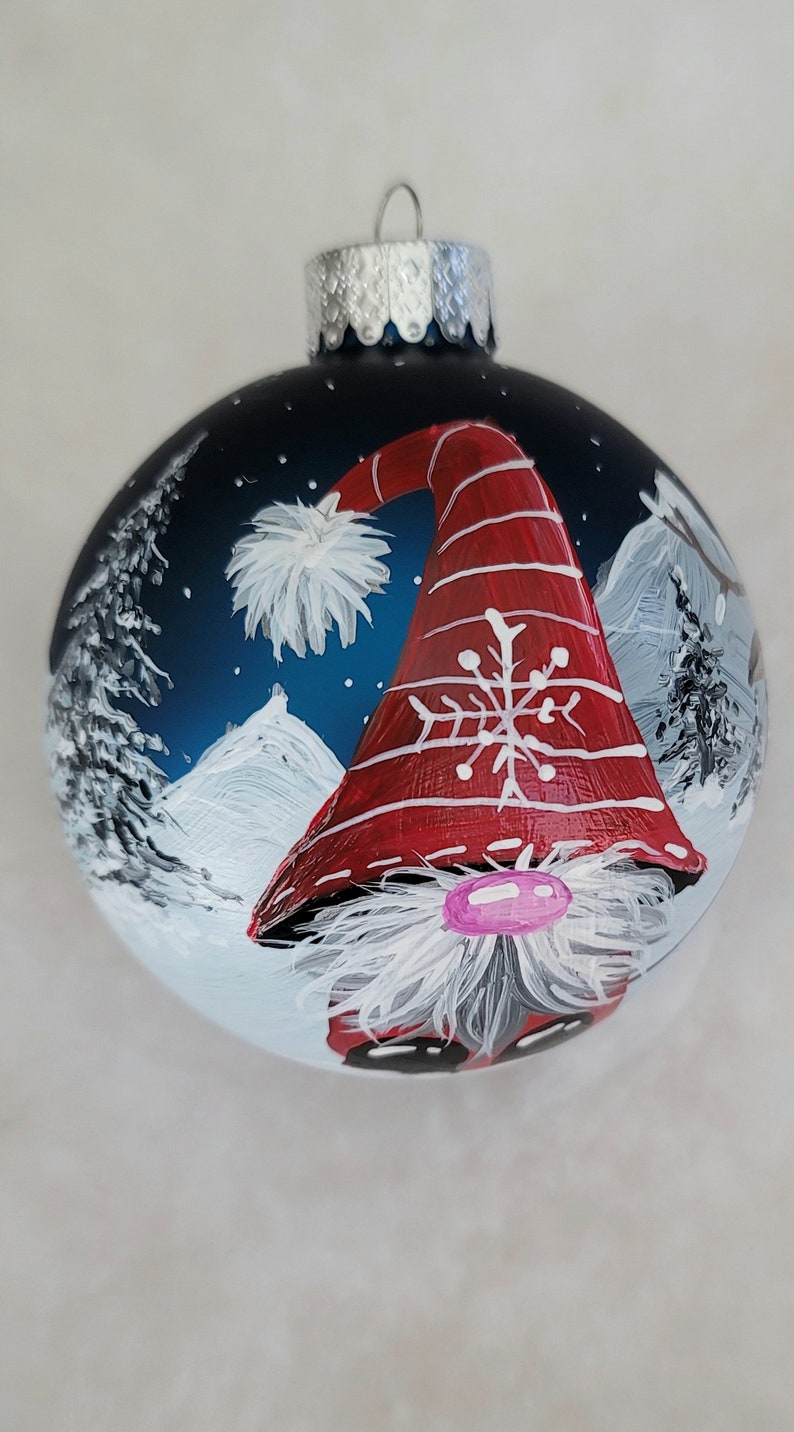 May include: A hand-painted Christmas ornament featuring a gnome with a red and white striped hat, a white beard, and a pink nose. The gnome is set against a blue background with white snow-capped mountains and white trees.