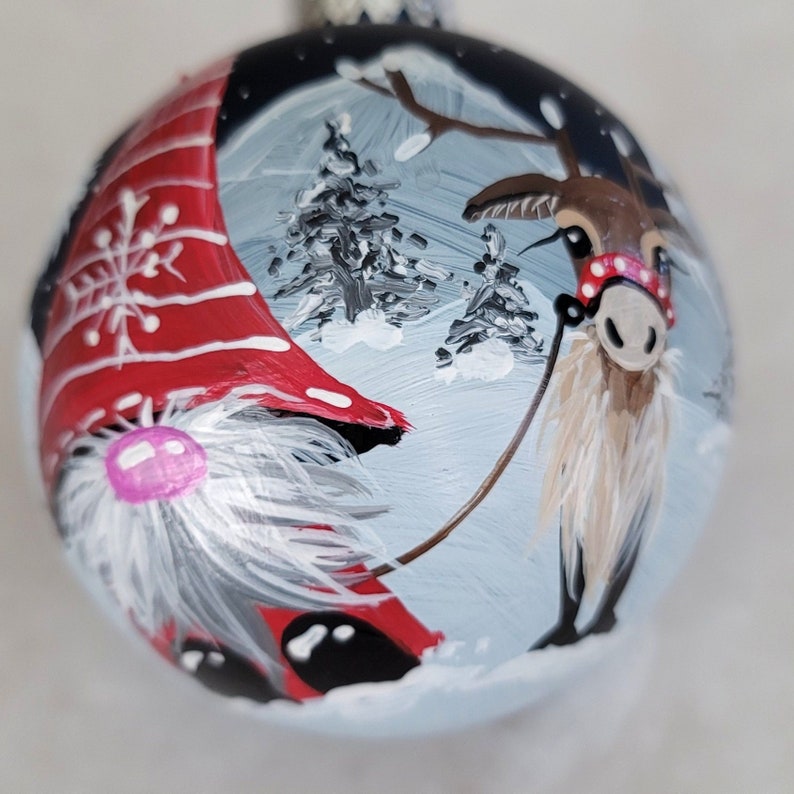 May include: A hand-painted Christmas ornament featuring a gnome and a reindeer in a snowy winter scene. The gnome wears a red hat with white trim and a white beard. The reindeer has a red and white striped nose and a white beard. The ornament is decorated with white snowflakes and trees.