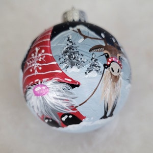 May include: A hand-painted Christmas ornament featuring a gnome wearing a red and white hat and a reindeer with a red and white scarf. The ornament is decorated with a winter scene, including snow-covered trees and a snowy background.