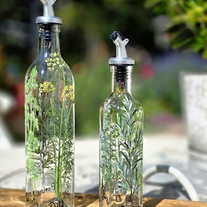 Può includere: Due bottiglie per olio in vetro trasparente con beccucci versatori color argento. La bottiglia più alta presenta piante di aneto dipinte a mano con fiori gialli e la parola Dill. La più piccola mostra rametti di rosmarino e la parola Rosemary.