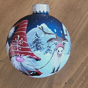 May include: A blue glass Christmas ornament with a hand-painted scene of a gnome and a reindeer in a snowy winter landscape. The gnome is wearing a red and white hat and the reindeer has a red and white collar.