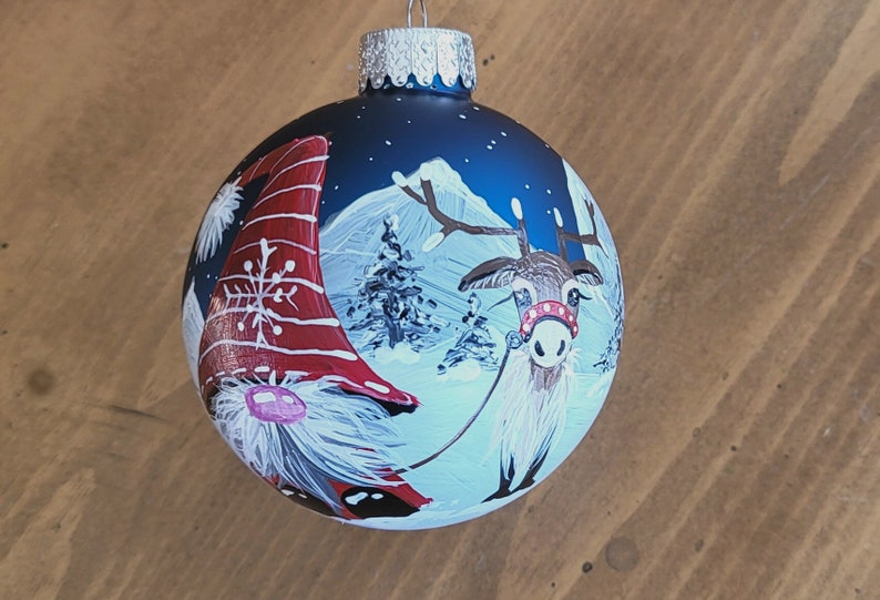 May include: A blue glass Christmas ornament with a hand-painted scene of a gnome and a reindeer in a snowy winter landscape. The gnome is wearing a red and white hat and the reindeer has a red and white collar.