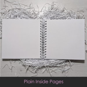 May include: An open, blank notebook with a spiral binding, resting on a bed of shredded silver paper. The notebook's pages are white, and the text "Plain Inside Pages" is at the bottom of the image.