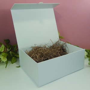 May include: A white gift box with a magnetic closure, open to reveal brown crinkle paper. The box is rectangular with a hinged lid, perfect for presenting gifts. The background is a soft pink with green and white floral accents.