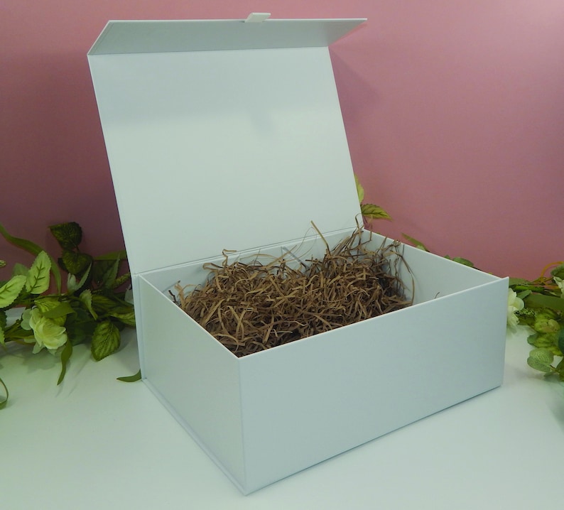 May include: A white gift box with a magnetic closure, open to reveal brown crinkle paper. The box is rectangular with a hinged lid, perfect for presenting gifts. The background is a soft pink with green and white floral accents.