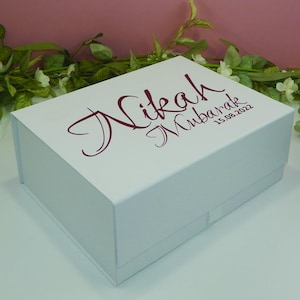 May include: A white gift box with a magnetic closure. The box has the words "Nikah Mubarak" and the date "15.08.2022" printed in a burgundy script font on the lid. The box is rectangular and sits on a white surface.