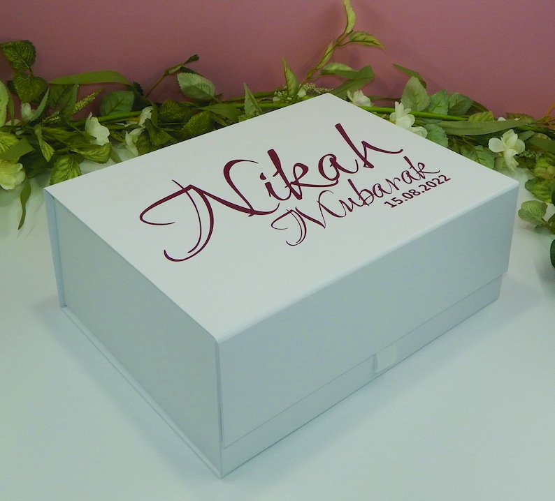 May include: A white gift box with a magnetic closure. The box has the words "Nikah Mubarak" and the date "15.08.2022" printed in a burgundy script font on the lid. The box is rectangular and sits on a white surface.