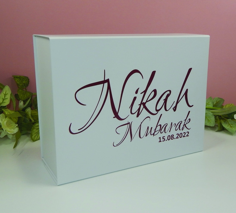 May include: A white gift box with a magnetic closure. The box features the words "Nikah Mubarak" in a burgundy script font, along with the date "15.08.2022". The box is a clean, elegant design.