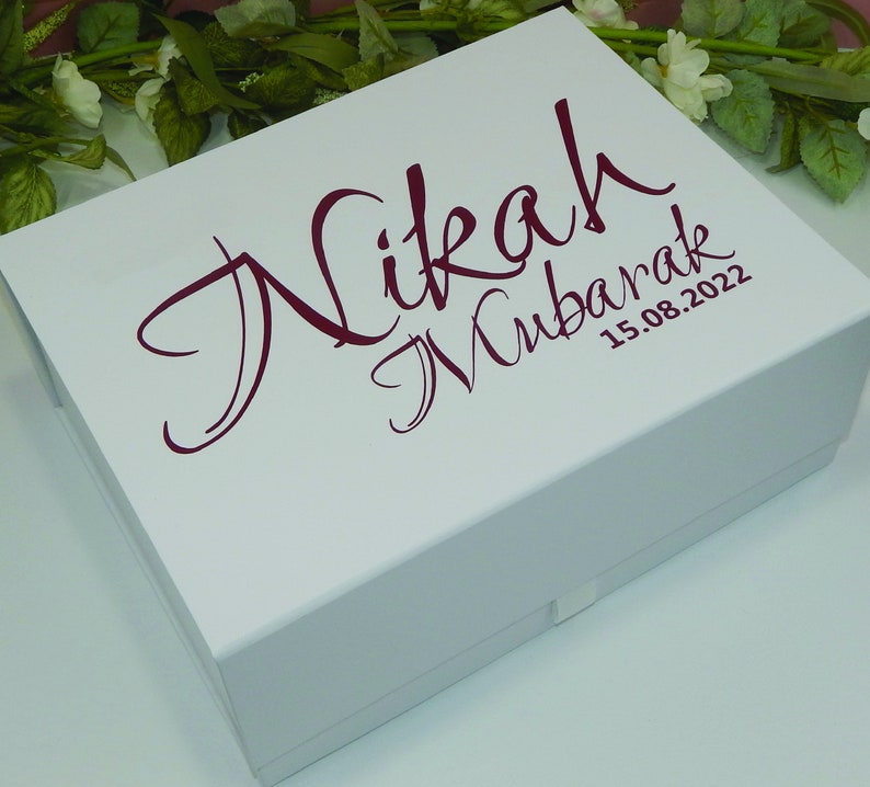 May include: A white gift box with a burgundy script that reads "Nikah Mubarak" and the date "15.08.2022". The box is rectangular with a lid and is surrounded by greenery and white flowers.