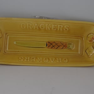 May include: A yellow ceramic cracker serving tray with a knife and crackers embossed on the surface. The tray reads "CRACKERS" on the top and bottom.